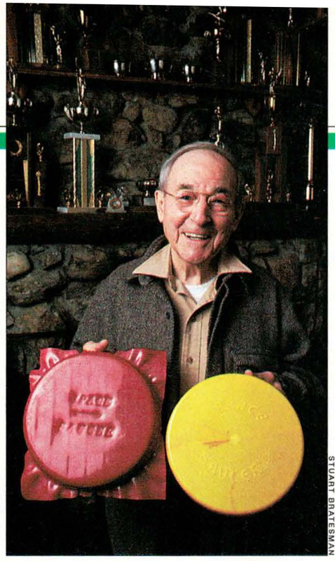 Ernest C. Bill Robes smiles while holding a red Space Saucer and a yellow Space Saucer in front of a trophy display.